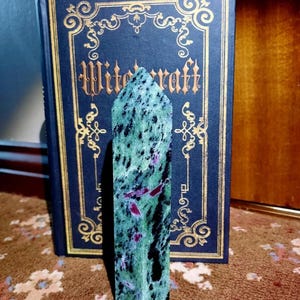 Może przedstawiać: Zielony i czarny szpic z kamienia szlachetnego z czerwonymi inkluzjami. Szpic stoi pionowo na wzorzystym dywanie przed książką zatytułowaną "Witchcraft".