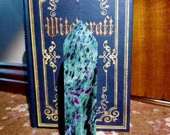 Op de afbeelding: Een groene en zwarte edelsteenpunt met rode insluitsels. De punt staat rechtop op een tapijt met een patroon voor een boek met de titel "Witchcraft".