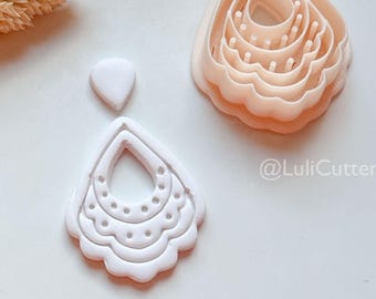 May include: Three light pink clay cutters in the shape of earrings and a small white clay piece are displayed on a white surface. One cutter is a layered teardrop shape, another is a single earring shape with a dotted design, and the third is a small oval.