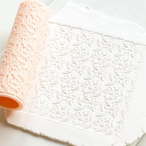 Può includere: Un mattarello color pesca chiaro con un motivo floreale accanto a un foglio di argilla bianca con lo stesso disegno in rilievo. Il mattarello è cilindrico e ha un interno arancione. Il foglio di argilla è rettangolare con una superficie strutturata.