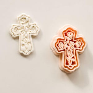 Könnte beinhalten: Zwei kreuzförmige Ausstecher. Einer ist weiß, der andere pfirsichfarben. Beide haben florale Designs. Das weiße Kreuz befindet sich links und das pfirsichfarbene Kreuz rechts. Der Hintergrund ist hell, cremefarben.