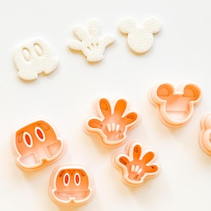 May include: A selection of light orange and white cookie cutters shaped like a cartoon mouse's face, hands, and shorts. The cutters are displayed on a white surface. The shapes are detailed and have a textured appearance.