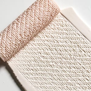 May include: A rectangular, off-white ceramic tray with a woven texture. The tray has a rolled-up section at one end, also with a woven pattern, and a raised border. The color is a soft, neutral tone, suitable for home decor.