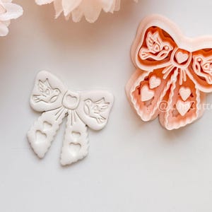 May include: Two bow-shaped cookie cutters. One is white, the other is peach-colored. Both have a bow design with heart and bird details. The peach-colored cutter has a visible outline. The cutters are on a white surface, with a peach-colored flower in the background.