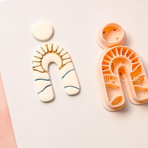 May include: A collection of polymer clay cutters and a finished clay piece. The cutters are peach-colored, featuring a sunburst design within an arch shape. The finished piece is white with gold and blue accents, also in an arch shape with a sunburst design.