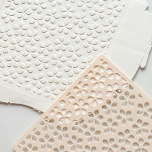 Peut inclure: Un rouleau à pâtisserie blanc est posé au-dessus d'une feuille d'argile blanche avec un motif floral. Un pochoir à motif floral de couleur pêche clair est à côté de l'argile. L'argile a été pressée avec le pochoir pour créer un motif floral.