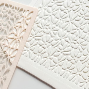 Op de afbeelding: Een close-up van twee knutselgereedschappen. Eén is een beige, opengewerkte sjabloon met een bloemenpatroon. De andere is een wit, rechthoekig stuk klei met een reliëf bloemmotief. De klei heeft een textuur, en de sjabloon ligt ernaast.