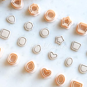 Op de afbeelding: Een verzameling polymeerklei uitsteekvormen in perzik- en beigetinten. De vormen omvatten vierkanten, cirkels, sterren en organische vormen zoals harten en druppels. De afbeelding bevat ook gedroogde bloemaccenten. Ideaal voor knutselen.