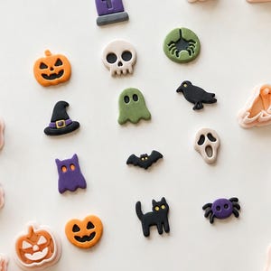 Op de afbeelding: Een verzameling kleine, kleurrijke Halloween-kleibedels en bijpassende lichtperzikkleurige plastic koekjesvormpjes. De bedels bevatten pompoenen, schedels, geesten, vleermuizen, katten, spinnen, raven en grafstenen in kleuren zoals oranje, zwart, paars en groen.