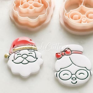 May include: A set of four Christmas cookie cutters. Two are plastic, depicting Santa Claus and Mrs. Claus faces. The other two are finished cookies, Santa and Mrs. Claus, with red and white details. Red berries are in the background.