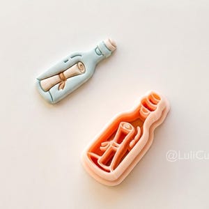May include: Two message-in-a-bottle cookie cutters. One is light blue with a scroll inside, the other is peach-colored and shows the scroll's outline. The cutters are on a white surface.