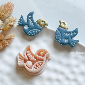 May include: Three bird-shaped items. Two are finished, blue with gold-coloured heads and white detailing. The third is a pink bird-shaped cookie cutter with floral and dot designs. The items are on a white surface.