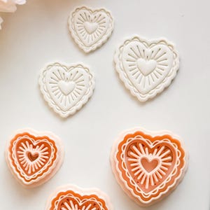 May include: A selection of heart-shaped cookie cutters in different sizes. The cutters are white and peach-coloured, featuring a scalloped edge and a sunburst design radiating from a heart in the centre. They are arranged on a white surface.