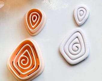 May include: A collection of polymer clay cutters in various sizes and shapes. The cutters are in the shape of abstract, organic forms with a spiral design. The colors are a warm peach and white. The cutters are arranged on a white surface.