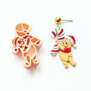 Könnte beinhalten: Ein pfirsichfarbener Kunststoff-Ausstecher in Form eines Bären mit Weihnachtsmütze und Zuckerstange, neben einem passenden Ohrring, der einen gelben Bären im roten Hemd mit Weihnachtsmütze zeigt, der eine rot-weiß-grüne Zuckerstange hält.