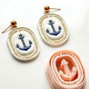 May include: Oval-shaped earrings with a nautical theme. Each earring features a blue anchor on a white background, framed by a gold-colored border. The earrings have gold-colored posts. A pink anchor-shaped mold is also present.