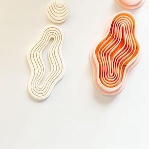 May include: A collection of polymer clay cutters in abstract organic shapes. The cutters are in shades of cream and peach, with layered, wavy designs. The shapes include a circle, a teardrop, and an irregular, flowing form, ideal for crafting.