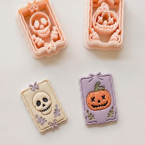 Può includere: Due tagliabiscotti in plastica color pesca chiaro a forma di cornici rettangolari con disegni di teschio e zucca, insieme a due pezzi di argilla in crema e lavanda con motivi in rilievo di teschio e zucca e dettagli floreali.