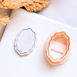 Op de afbeelding: Twee ovale koekjessnijders. Eén is zilverkleurig met een textuur aan de binnenkant, en de andere is perzikkleurig met een gelaagd ontwerp. Gedroogde tarwearen zijn op de achtergrond. De snijders liggen op een wit oppervlak.