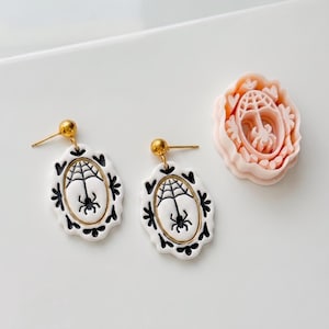 May include: A pair of oval-shaped earrings featuring a white base with black spider and web illustrations, framed by a scalloped edge with black heart accents and gold-tone posts, accompanied by a light pink plastic oval-shaped clay cutter.