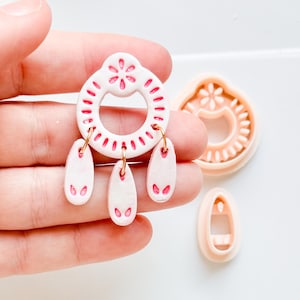 May include: Handmade white and pink earrings with a floral design. The earrings feature a circular top with a flower and three teardrop pendants. Additional pink clay cutters are also visible in the image.