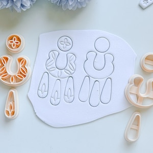 May include: A collection of polymer clay earring cutters and a clay slab with earring designs. The cutters are in various shapes, including a flower, a U-shape, and teardrops. The clay slab has two earring outlines. The colors are white and peach.