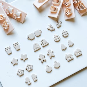 Könnte beinhalten: Eine Sammlung hellbeige Ausstecher in verschiedenen Formen, darunter Sterne, Häuser, Lebkuchenmänner und Ornamente. Einige Ausstecher sind an rosa Schlüsselanhängern befestigt. Die Ausstecher liegen auf einer weißen Oberfläche.