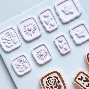 Puede incluir: Colección de sellos cuadrados rosa claro con varios diseños, incluyendo rosas, soles, corazones y pájaros. Los sellos están dispuestos sobre una superficie blanca, mostrando sus detallados patrones en relieve. Ideales para manualidades y proyectos decorativos.