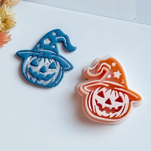 May include: Two Halloween-themed pumpkin and witch hat shaped decorations. One is blue with a silver pumpkin face and the other is orange with a pink pumpkin face. The decorations are on a white surface.