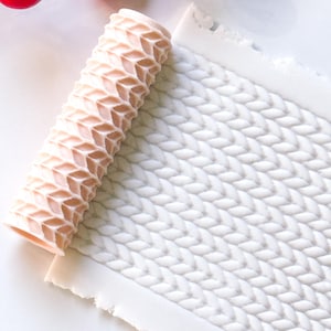 May include: A light pink textured rolling pin next to a white clay sheet with a knitted pattern. A sprig of red berries is in the background. The rolling pin is designed to create a knitted texture on clay or dough.