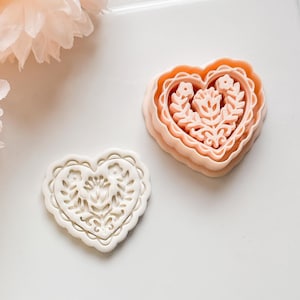 May include: Two heart-shaped cookie cutters. One is a light peach color, and the other is white. Both have a floral design in the center. The peach-colored cutter is a three-piece set. The white cutter is a single piece.