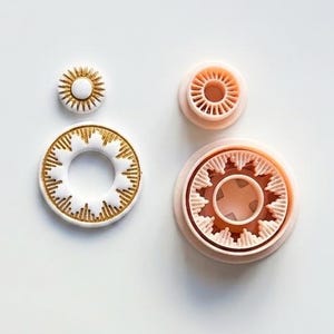 May include: Two white circular clay components featuring gold sunburst patterns, paired with two light peach-coloured plastic cutters. The smaller circular component and its corresponding cutter are positioned above the larger circular component and its cutter.