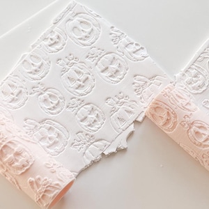 May include: A light pink cylindrical rolling pin with a repeating pumpkin pattern used to emboss white clay. The tool creates a textured surface on the clay, and several small plastic cookie cutters with pumpkin and spiderweb designs are visible.