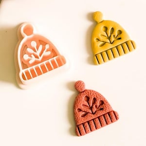 Op de afbeelding: Drie decoratieve wintermutenmagneten in tinten oranje, geel en rood. Elke muts heeft een sneeuwvlokontwerp en een geribbelde rand. Een rode bol is ook zichtbaar.