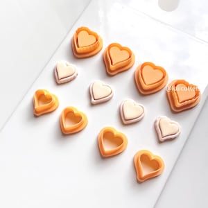 May include: A collection of heart-shaped cookie cutters in various sizes and designs. The cutters are in shades of orange and cream, with some featuring a layered design. The image is taken from a slightly elevated angle, showcasing the cutters against a white background.
