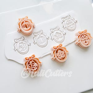 May include: A set of peach-colored plastic cookie cutters featuring oval frames with bows, containing designs of a pumpkin, a squirrel, a maple leaf, and a mushroom. The cutters are shown pressing into white clay, leaving detailed impressions.