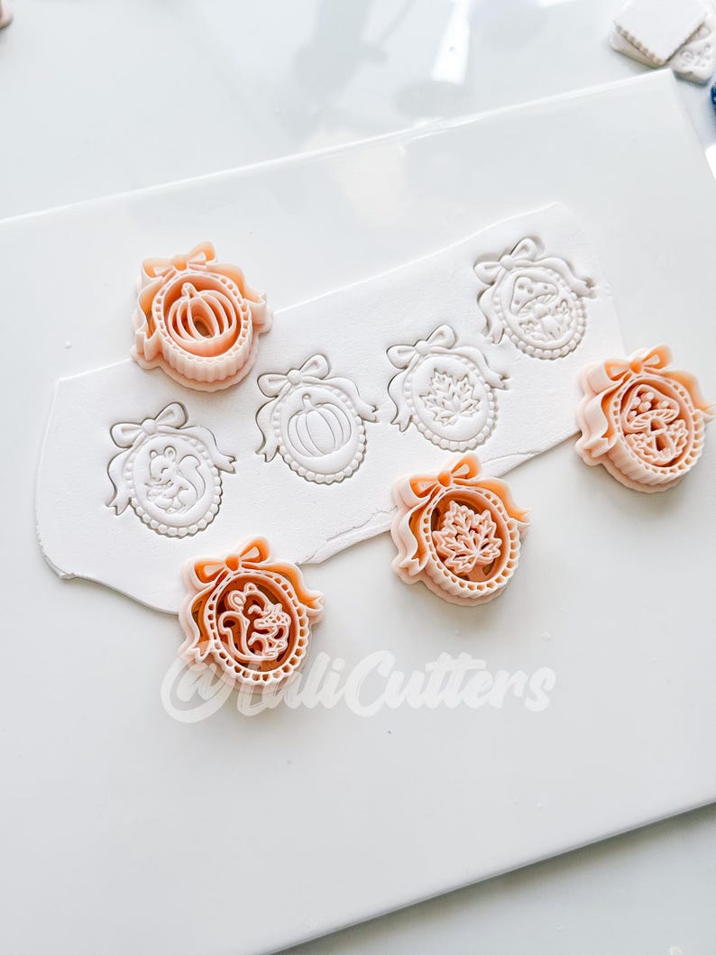 May include: A set of peach-colored plastic cookie cutters featuring oval frames with bows, containing designs of a pumpkin, a squirrel, a maple leaf, and a mushroom. The cutters are shown pressing into white clay, leaving detailed impressions.