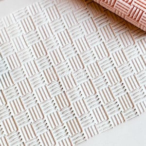 May include: A light pink rolling pin with a basket weave design sits above a piece of white clay, also imprinted with the same pattern. The image is a close-up, showing the texture of the rolling pin and clay. The rolling pin is used for crafts.