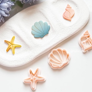 Può includere: Un set di cinque stampini per biscotti a forma di conchiglie e stelle marine. Gli stampini sono in plastica rosa e sono disposti su una superficie bianca. Gli stampini sono perfetti per fare biscotti, fondant o altre decorazioni.