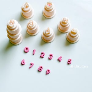 May include: A selection of light pink number and letter cookie cutters. The cutters are stacked and arranged on a light blue surface. The numbers 0-9 are also displayed in a smaller size. The text "@lulicutters" is visible.