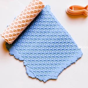 Può includere: Un mattarello strutturato arancione chiaro e un foglio di argilla blu con un motivo a scaglie. È presente anche un piccolo tagliapasta a forma di pesce. Gli strumenti sono probabilmente per l'artigianato o la cottura.