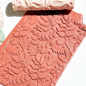 May include: A terracotta-colored, leaf-patterned roller and a matching sheet. The roller is cylindrical with a raised floral design. The sheet has a similar embossed pattern of leaves, berries, and vines. The items are likely used for crafts or decorative purposes.