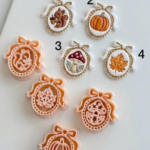 May include: A collection of oval-shaped cookie cutters and corresponding embossed cookies featuring autumn themes. The designs include a squirrel with an acorn, a pumpkin, a mushroom, and a maple leaf, all framed by a decorative bow and border.