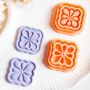 May include: Two sets of square cookie cutters with a flower design. One set is orange and the other is purple.