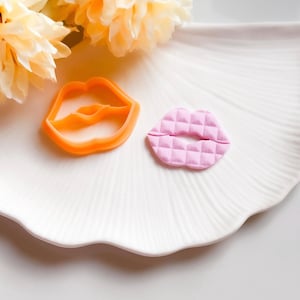 Pode incluir: Um cortador de biscoitos de plástico laranja em forma de lábios ao lado de uma decoração rosa acolchoada em forma de lábios. Os itens estão em um prato branco com bordas onduladas, com flores amarelas ao fundo. O cortador é um contorno sólido.