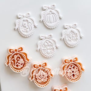 May include: A collection of oval-shaped cookie cutters and their corresponding white embossed dough impressions. The designs feature a squirrel, a pumpkin, a mushroom, and a maple leaf, each framed by a decorative border with a bow at the top.