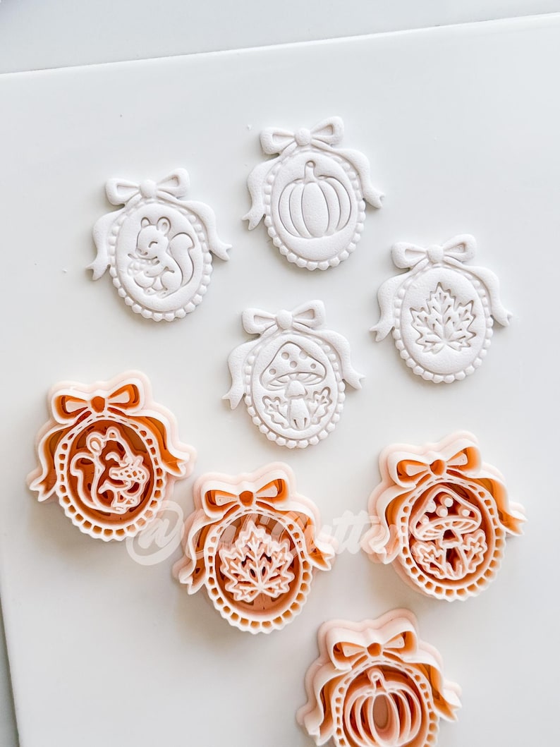 May include: A collection of oval-shaped cookie cutters and their corresponding white embossed dough impressions. The designs feature a squirrel, a pumpkin, a mushroom, and a maple leaf, each framed by a decorative border with a bow at the top.