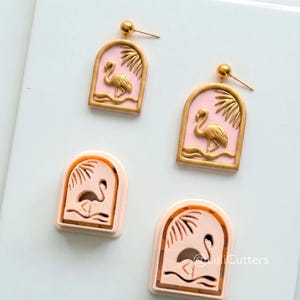 Peut inclure: Boucles d'oreilles et emporte-pièces assortis avec des flamants roses roses et dorés. Les boucles d'oreilles présentent un flamant rose doré et un motif de feuilles de palmier dans une arche rose. Les emporte-pièces ont un design similaire.