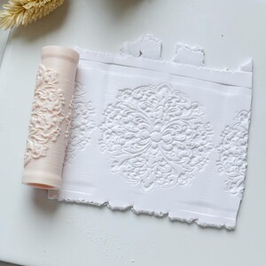 May include: A light pink roller with a floral design next to a white sheet with an embossed floral pattern. The roller is cylindrical and has a detailed, raised floral pattern. The white sheet shows the embossed design, creating a textured effect.