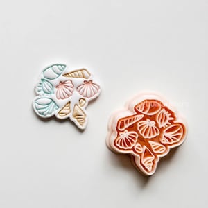 Könnte beinhalten: Zwei muschelförmige Ausstecher. Einer ist hellrosa, der andere weiß mit blauen, rosa und goldenen Akzenten. Die Ausstecher sind mit verschiedenen Muschelformen gestaltet, perfekt für Leckereien zum Thema Strand.